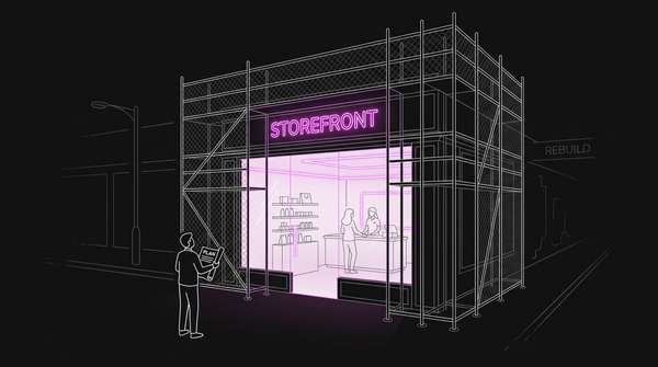 A shopfront still lit and trading at night while its outer facade stands stripped back inside scaffolding, with one person outside holding a rolled plan and looking up
